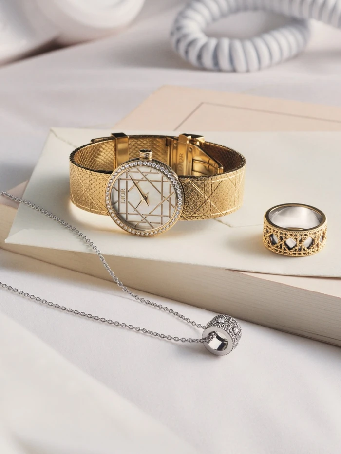 Gold Dior wristwatch with a diamond-paved bezel, matching gold ring, and silver pendant necklace on pale paper