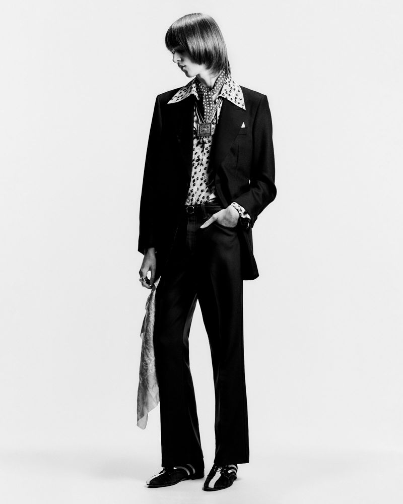 A stylish young adult man in a black suit with a patterned shirt and scarf, posing in a studio with a confident expression.