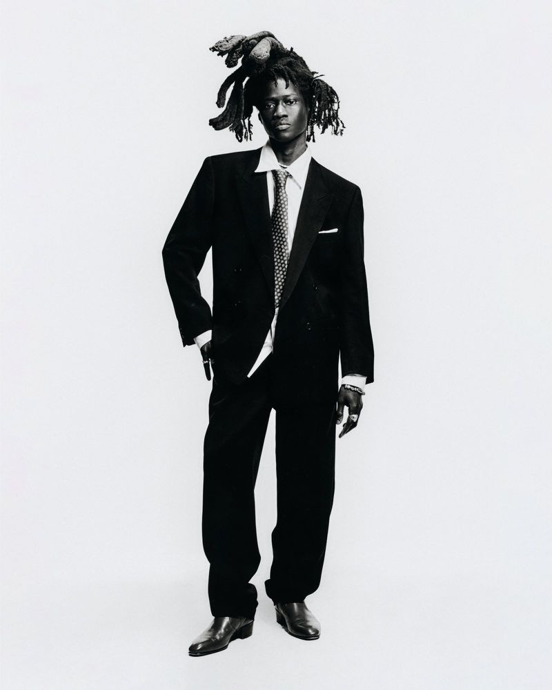 Monochrome portrait of Black male in a black suit, patterned tie, and unique headwear, posing in a photo studio against a white backdrop.