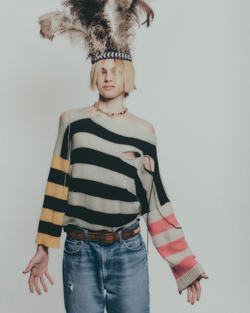 Person in a striped sweater and feather headdress poses in a studio. Fashionable street style with light denim jeans and jewelry.