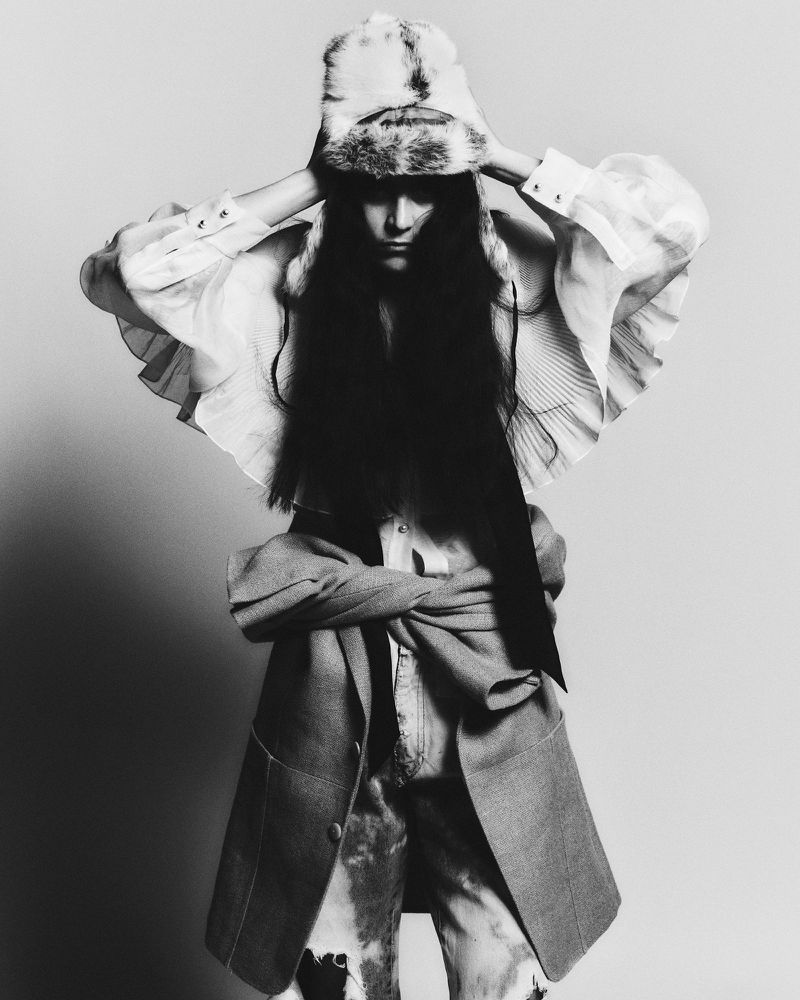 Monochrome fashion shot of a woman in a fur hat, layered shirt, tie-dye jeans, and coat, standing in front of a plain backdrop.
