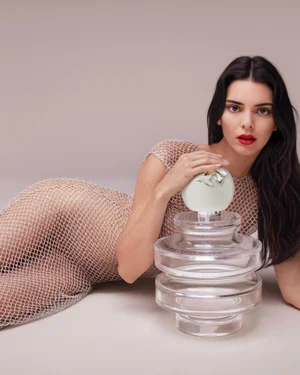 Kendall Jenner lying down wearing a sheer mesh dress beside a large perfume bottle against a neutral studio background