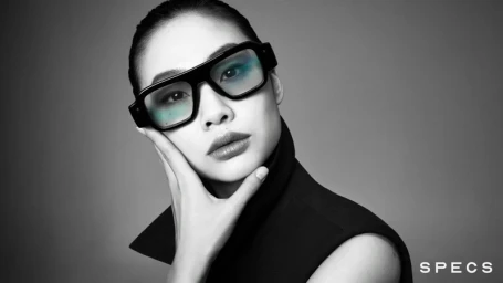 Hoyeon Jung wearing chunky black smart glasses and a dark high-collar top against a plain grey studio backdrop by Steven Meisel.