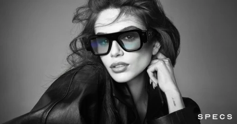 Black and white portrait of a woman wearing thick black glasses and a leather jacket against a seamless background.