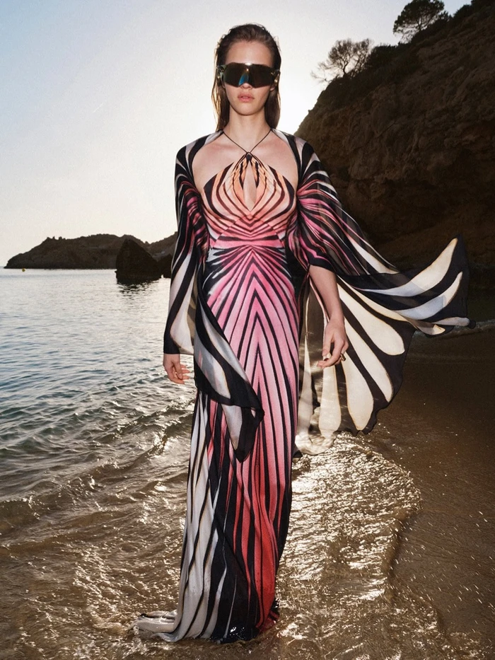 Model wearing a patterned maxi dress and oversized sunglasses standing on a sandy beach by the ocean.