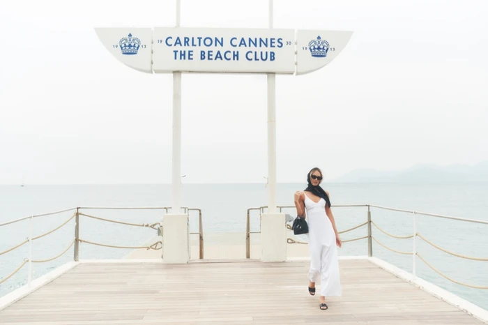 Model Candice Swanepoel walking on a wooden pier beneath the Carlton Cannes Beach Club sign for Alo