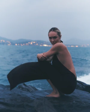 Model Candice Swanepoel crouching on dark rocks by the ocean at dusk for Alo Summer Atelier 2026.