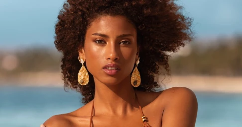 Close-up portrait of model Imaan Hammam wearing large gold statement earrings against an out-of-focus sandy beach backdrop.