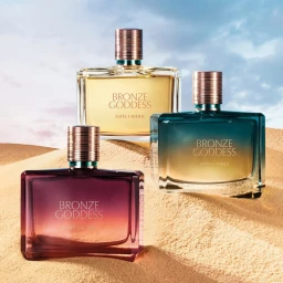 Three glass perfume bottles of Bronze Goddess by Estee Lauder set on sand dunes against a blue sky with soft clouds.