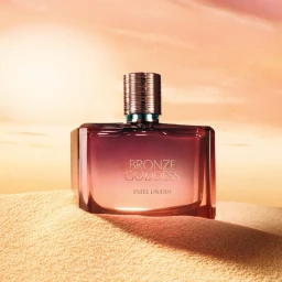 Estee Lauder Bronze Goddess perfume bottle resting in sand against a warm sunset sky background.