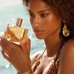 Imaan Hammam holding an Estee Lauder Bronze Goddess perfume bottle outdoors near water with warm sunlight.