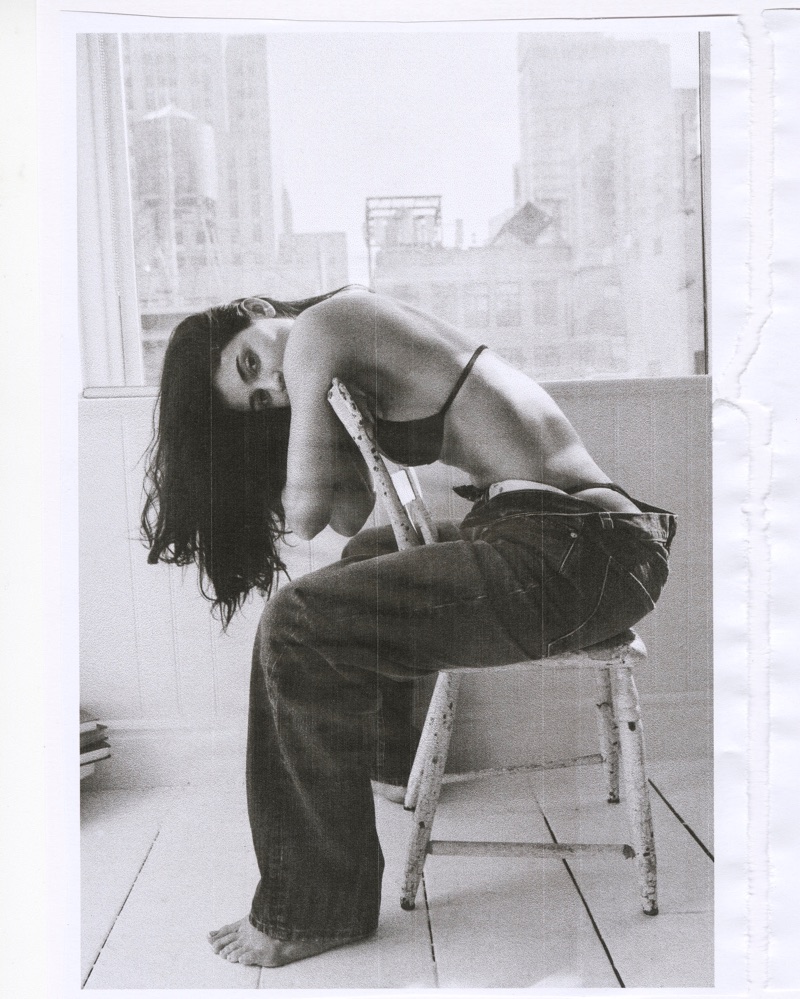 Kylie Jenner seated backwards on a white chair wearing loose jeans and a black bikini top in a light-filled room with city window views