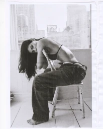 Kylie Jenner seated backwards on a white chair wearing loose jeans and a black bikini top in a light-filled room with city window views