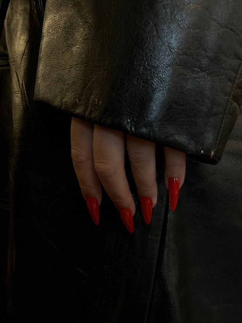 Close-up on a manicured hand with bright red stiletto nails resting on a black leather jacket. Focus on nail art and fashion.