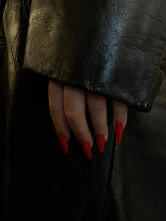 Close-up on a manicured hand with bright red stiletto nails resting on a black leather jacket. Focus on nail art and fashion.