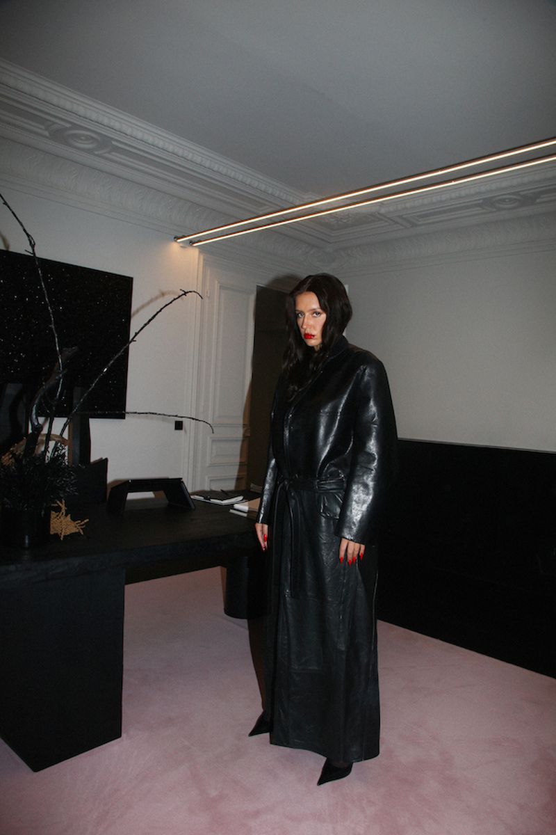 Brunette woman with red lipstick poses indoors wearing a full-length black leather coat, standing on a pink carpet against a white wall.