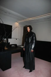 Adele Exarchopoulos wearing a full-length black leather trench coat standing on pink carpeting in a minimalist room.