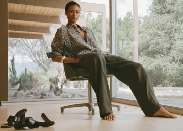 Model seated in an office chair wearing a patterned button-down shirt and trousers inside a modern home with large windows.
