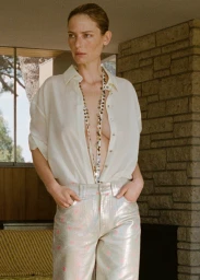 Woman wearing an unbuttoned white shirt and patterned metallic trousers standing indoors by a stone wall.