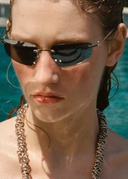 Close-up portrait of a woman wearing dark rimless sunglasses and a chunky gold chain necklace by a pool