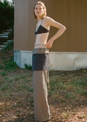 Young woman in a black bralette and layered trousers standing outdoors near a building exterior.