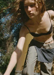 Model with wavy brown hair wearing a black spaghetti strap top and green trousers outdoors in sunlight