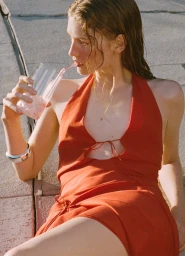 Woman in a red halter dress drinking from a glass with a straw while seated outdoors on concrete pavement.