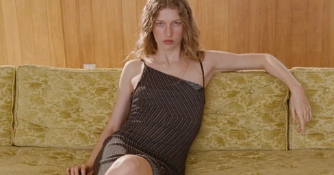 Model wearing a dark striped dress reclining on a patterned green couch against wood-paneled walls.