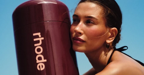 Hailey Bieber presses her cheek against a large glossy burgundy Rhode product container under a bright blue sky.