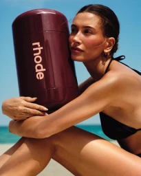 Hailey Bieber holding a large branded burgundy container on a beach for Rhode Pocket Bronze campaign