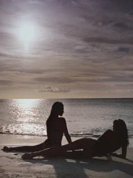 Two models silhouetted on a sandy beach during sunset, with ocean waves gentle in the background under a cloudy sky.