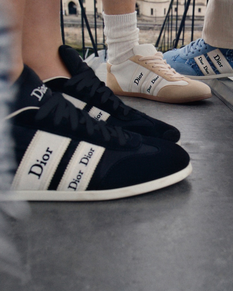 Close-up of low-top Dior ribbon sneakers in black, off-white, and blue denim worn by models standing outdoors.