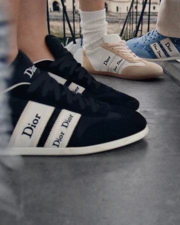 Close-up of low-top Dior ribbon sneakers in black, off-white, and blue denim worn by models standing outdoors.