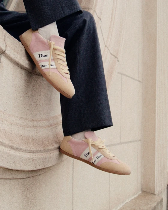 Close-up of Dior pink and beige sneakers on feet with dark trousers against a stone architectural background.