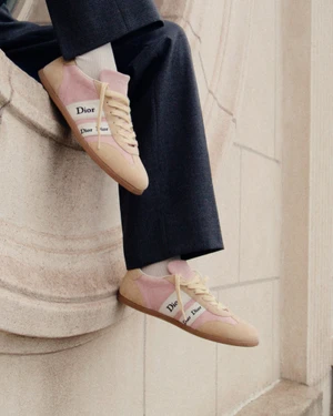 Close-up of Dior pink and beige sneakers on feet with dark trousers against a stone architectural background.
