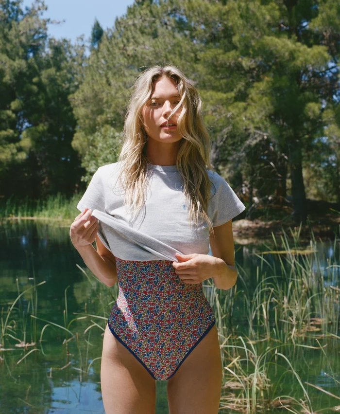Martha Hunt wearing a grey t-shirt and floral swimsuit standing outdoors by a calm water body with trees.