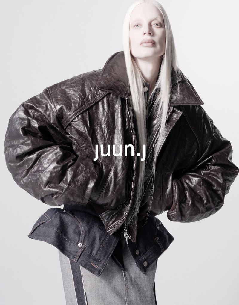 Fashion shot of a person with long white hair, wearing a dark brown leather jacket and dark and light blue denim, against white backdrop.