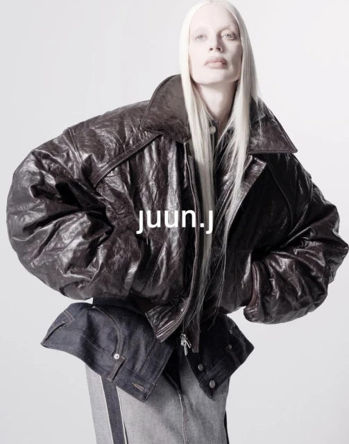 Fashion shot of a person with long white hair, wearing a dark brown leather jacket and dark and light blue denim, against white backdrop.