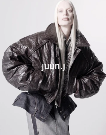 Fashion shot of a person with long white hair, wearing a dark brown leather jacket and dark and light blue denim, against white backdrop.
