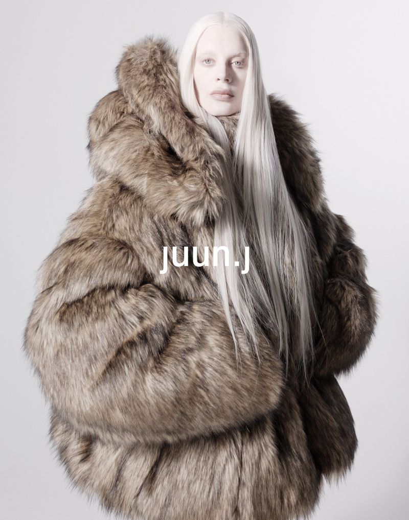 Model Kristen McMenamy wearing an oversized brown faux-fur hooded coat with long platinum hair against a white background.