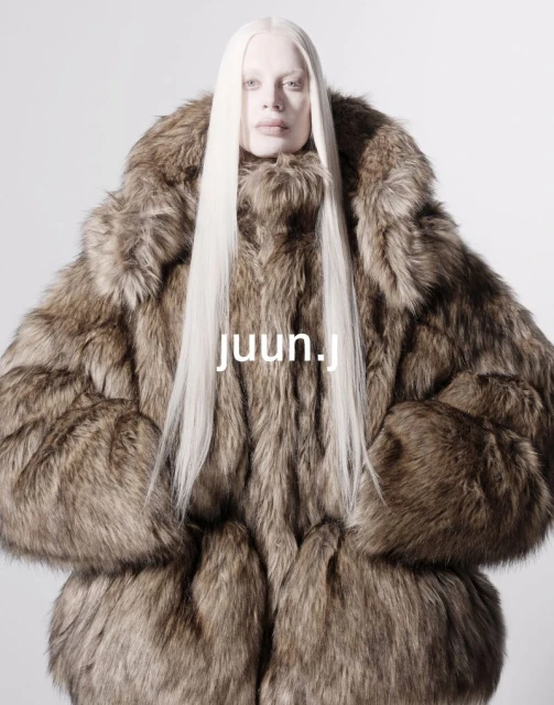 Fashion portrait of a person wearing a large fur coat and long white hair, set against a light grey background. The focus is on the coat.