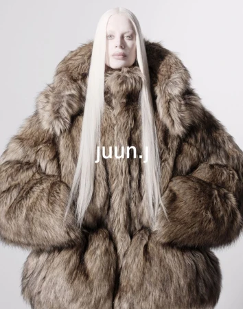Fashion portrait of a person wearing a large fur coat and long white hair, set against a light grey background. The focus is on the coat.