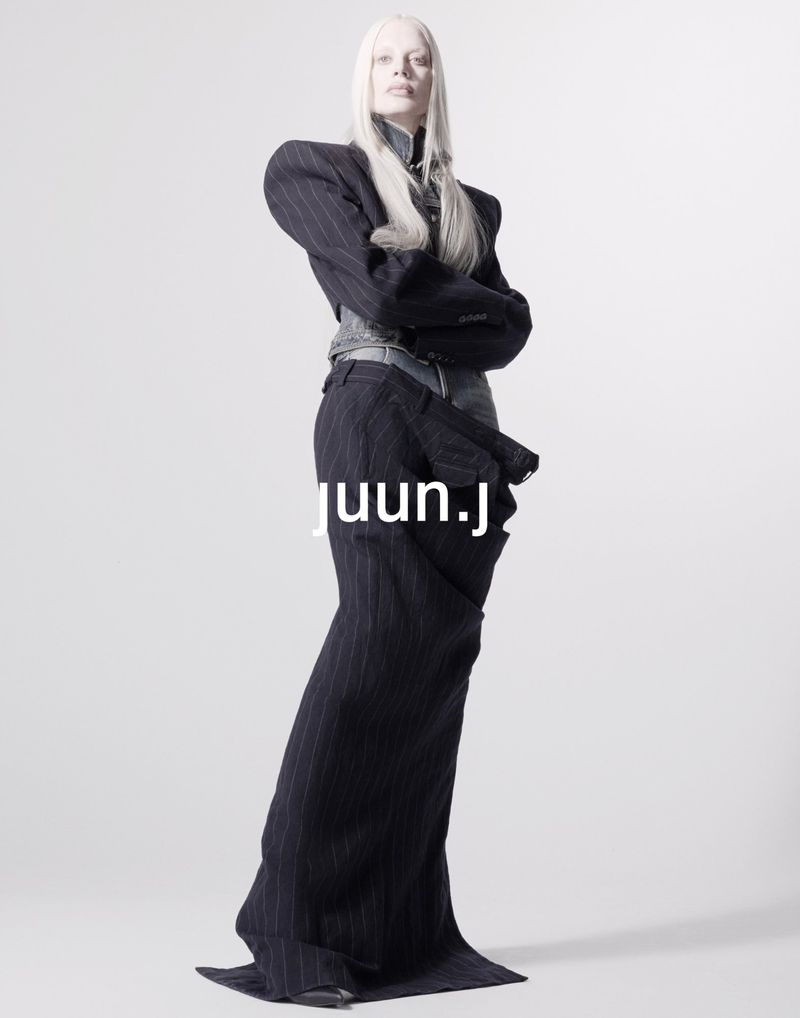 Avant-garde fashion shot of a pale woman in a pinstripe suit with exaggerated shoulders and a long skirt, posing against a white backdrop.