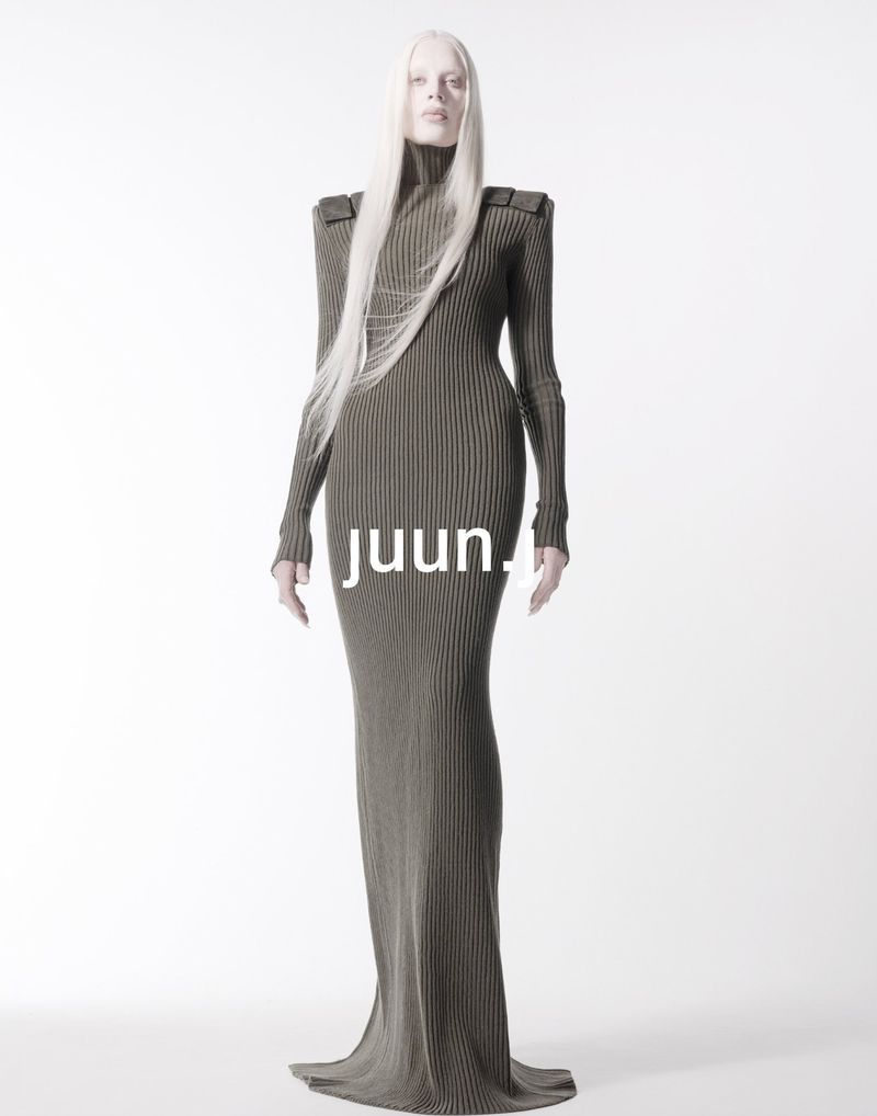 Fashion model with long silver hair poses in a full-length ribbed olive-green dress against a bright white backdrop in a studio setting.