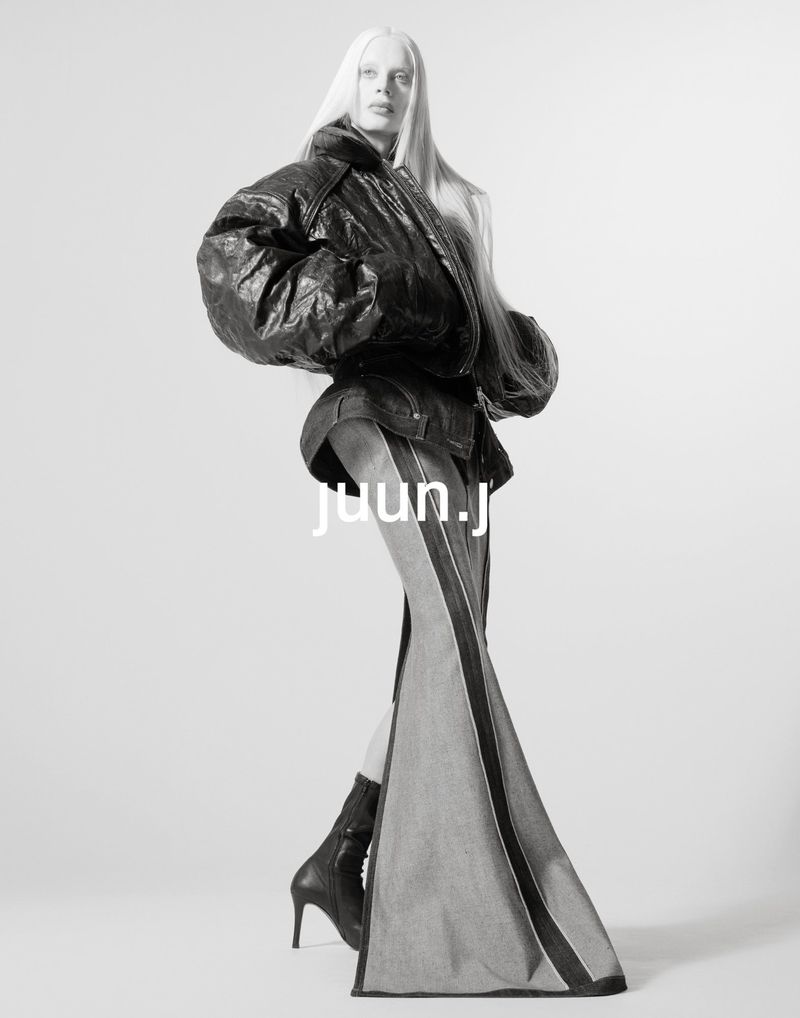 Monochrome fashion shot of a woman with platinum hair wearing a black leather jacket, denim skirt, black heeled boots in a photo studio.