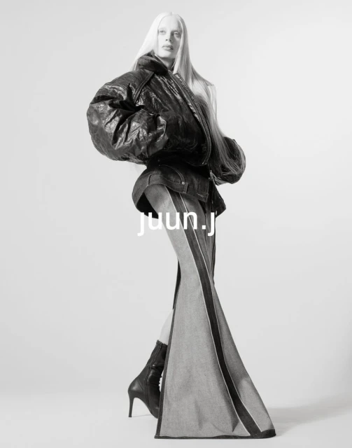 Monochrome fashion shot of a woman with platinum hair wearing a black leather jacket, denim skirt, black heeled boots in a photo studio.