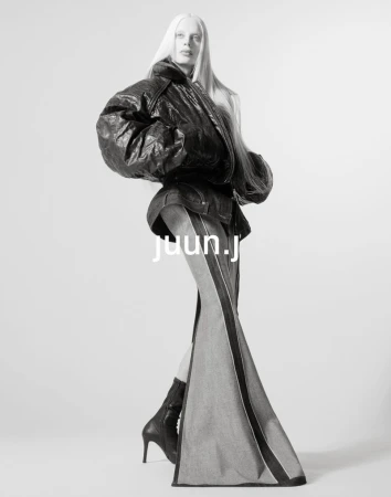 Monochrome fashion shot of a woman with platinum hair wearing a black leather jacket, denim skirt, black heeled boots in a photo studio.