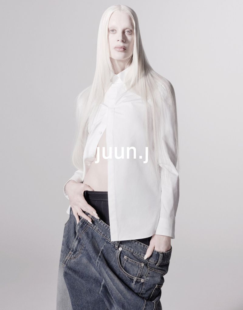 Fashion shot of androgynous model with long white hair, wearing a white button-down shirt and dark denim pants against a white backdrop.