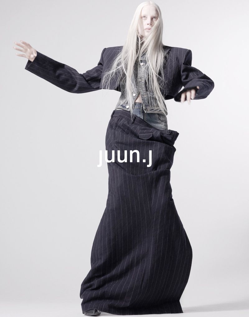 Fashion model poses in studio wearing a pinstripe blazer and floor-length skirt with denim shirt against a plain white backdrop.