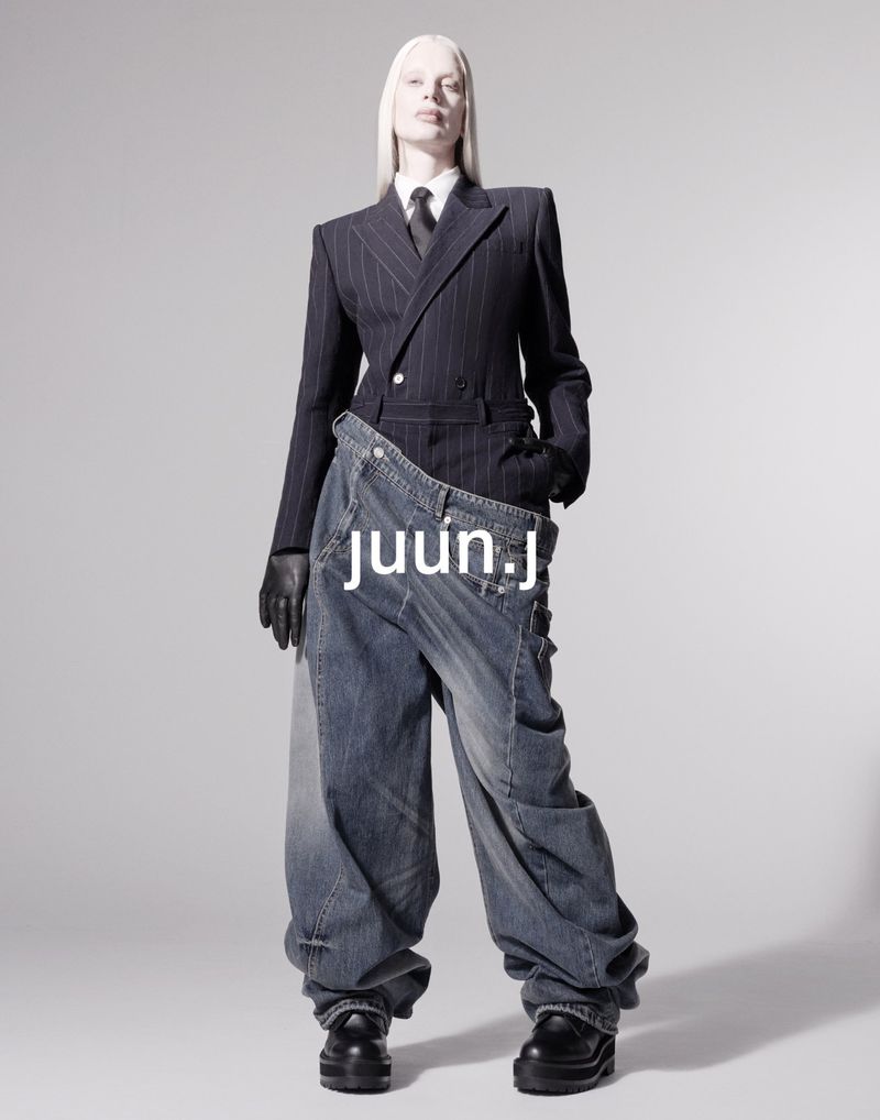 Fashion shoot of a slender adult woman with white hair wearing a dark suit and oversized jeans in a photo studio against a gray background.